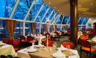 a well - lit restaurant with multiple dining tables , each set for a meal , and large windows offering views of trees outside at Achat Hotel Kaiserhof Landshut