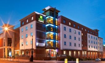 a large hotel with a green sign on the front , surrounded by trees and other buildings at Holiday Inn Express Stevenage