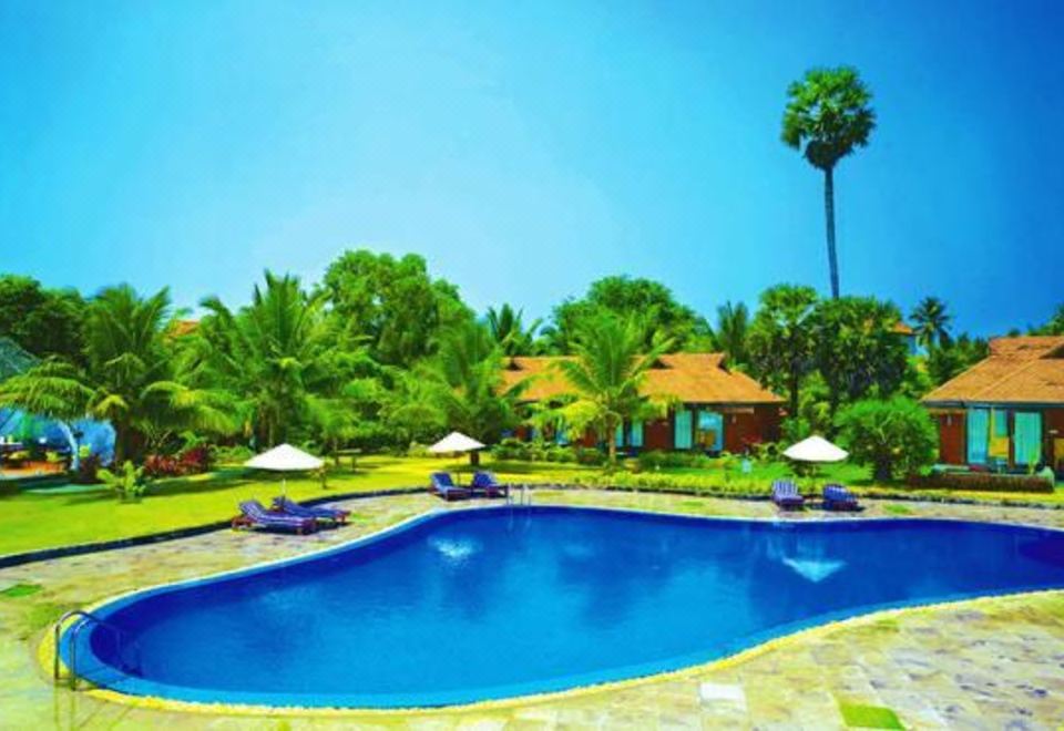 a beautiful swimming pool with umbrellas and lounge chairs , surrounded by lush greenery and clear blue skies at Poovar Island Resort