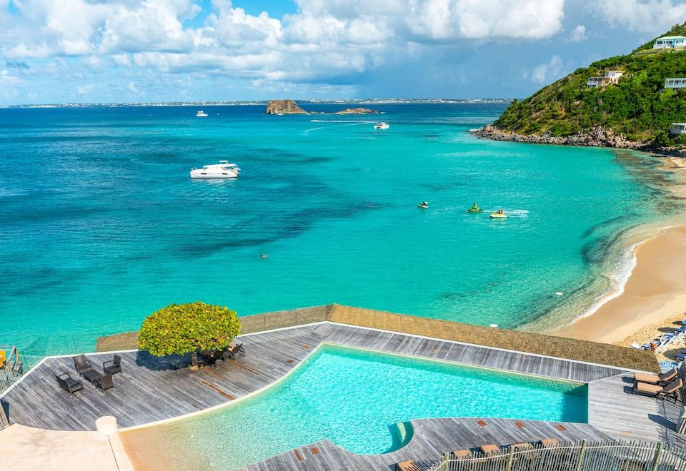 a beautiful beach scene with a pool surrounded by palm trees , a dock , and a boat on the water at Grand Case Beach Club