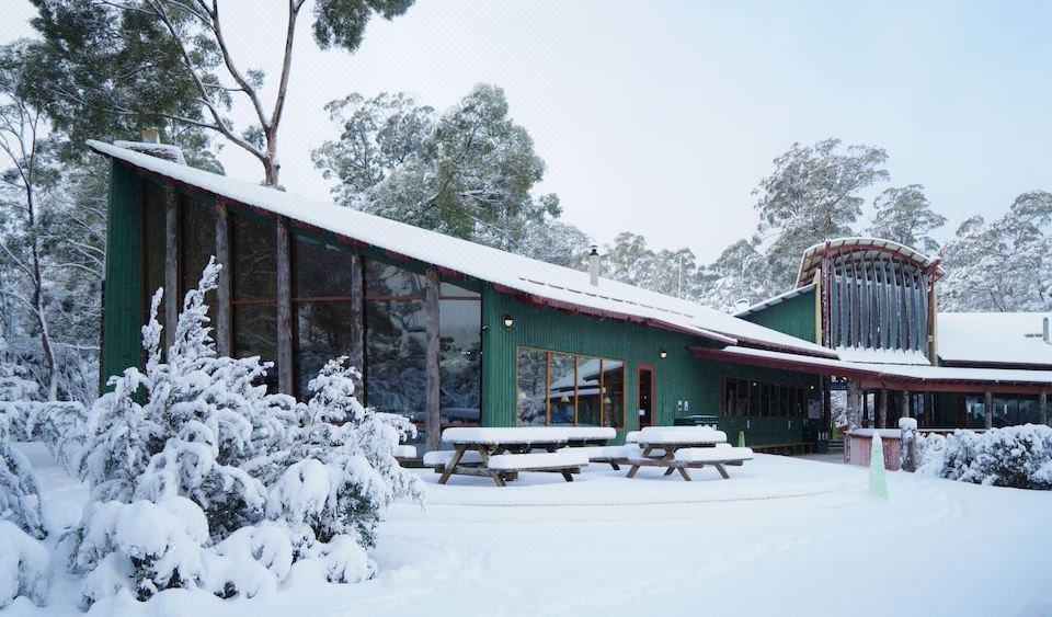 a large green building with a covered walkway and picnic tables is surrounded by snow at Lake St Clair Lodge