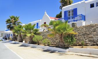 a white house with blue shutters is surrounded by palm trees and palm - lined walkways , set against a clear blue sky at Mykonos Beach Hotel