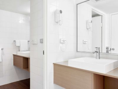 a bathroom with white walls , wooden furniture , and a large mirror above the sink , as well as a hairdryer hanging on the wall at Ibis Melbourne Hotel and Apartments