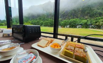 a table with a plate of food , including sandwiches and fruit , is set up in front of a window overlooking a field at Phuphayot Resort