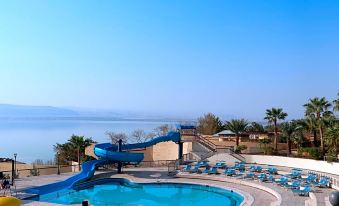 a large swimming pool with a water slide is surrounded by lounge chairs and palm trees at Dead Sea Spa Hotel