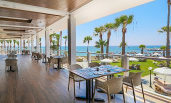 a large dining area with wooden tables and chairs , overlooking the ocean , with palm trees in the background at Golden Bay Beach Hotel