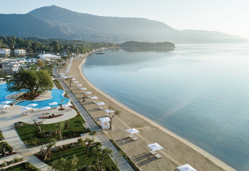 a beautiful beach with clear blue water , white umbrellas , and sun loungers , surrounded by mountains and trees at Ikos Dassia