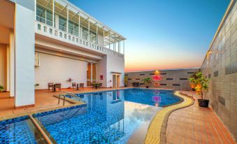 a large , blue - tiled swimming pool is surrounded by lounge chairs and tables , with a white building in the background at favehotel Bitung