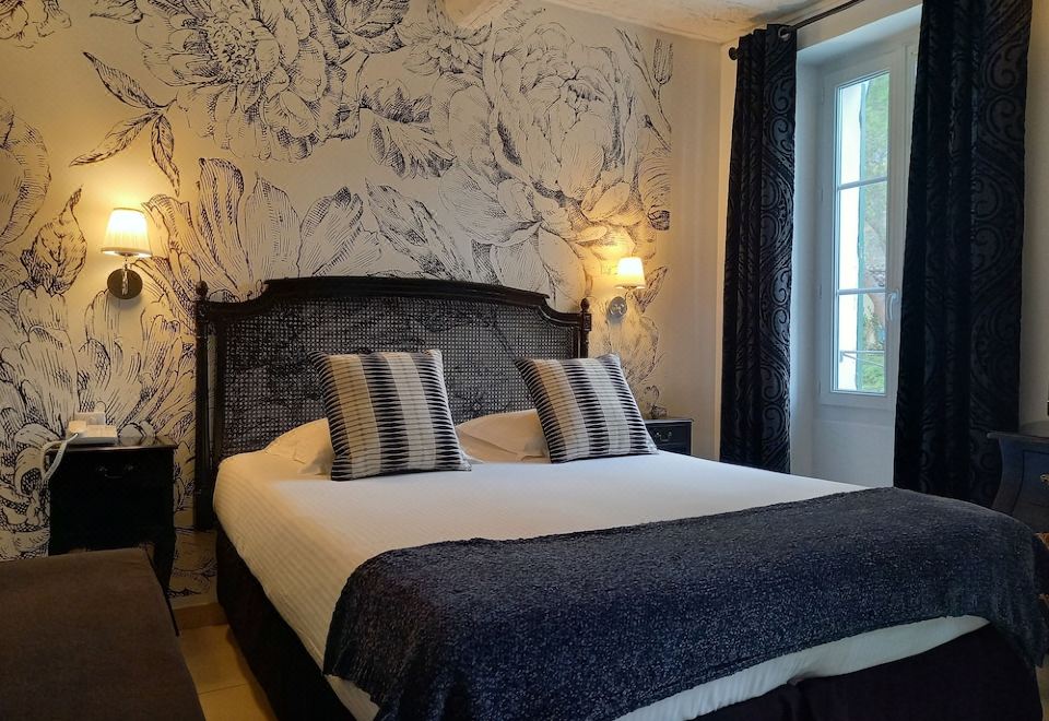 a large bed with a black headboard and white linens is situated in a room with floral wallpaper , two lamps on either side of the at Hôtel Restaurant Notre Dame