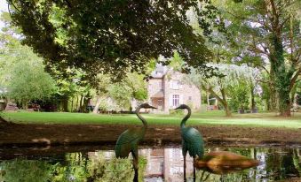 two green flamingos standing in a pond with a house and trees in the background at La Maison Rouge Broceliande