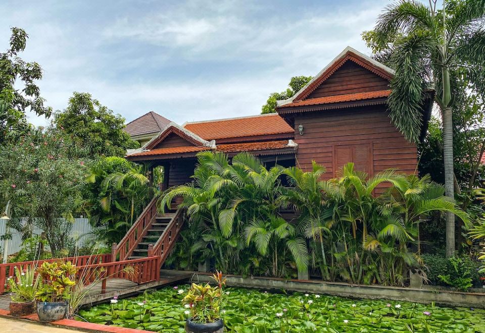 a red - roofed building surrounded by lush greenery and a pond with lily pads , with stairs leading up to the entrance at Vimean Sovannaphoum Resort