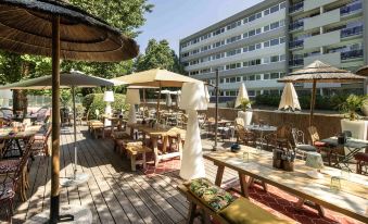 a wooden deck with umbrellas , tables , and chairs set up for outdoor dining , surrounded by trees and buildings at Novotel Avignon Nord