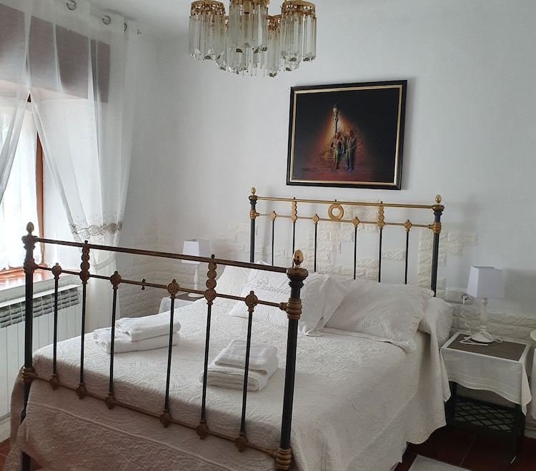 a white bedroom with a metal bed , curtains , and a painting on the wall , under a chandelier at Casa Rural la Plazuela de Mari