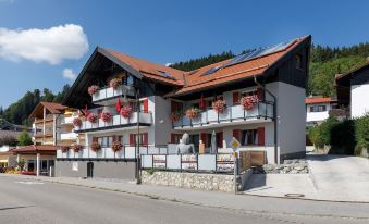 a large , multi - story building with a red roof and balconies is surrounded by mountains and has flowers on the balconies at Hotel Haus am See