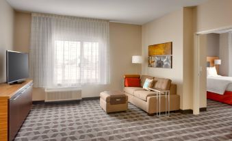 a living room with a brown couch , ottoman , and a painting on the wall , along with a window and lamp at TownePlace Suites Clovis