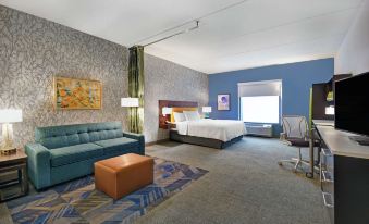 a modern hotel room with a blue couch , wooden desk , and white bed , along with a desk , chair , and rug at Home2 Suites by Hilton Lawrenceville Atlanta Sugarloaf