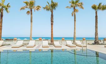 a long , empty pool surrounded by white lounge chairs and palm trees , with the ocean in the background at Grecotel Plaza Beach House