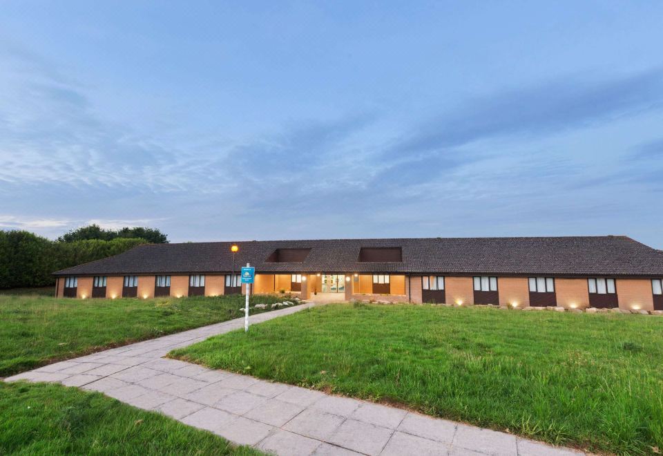 a large , modern building with multiple entrances and a grassy field in front of it at Days Inn by Wyndham Sutton Scotney South