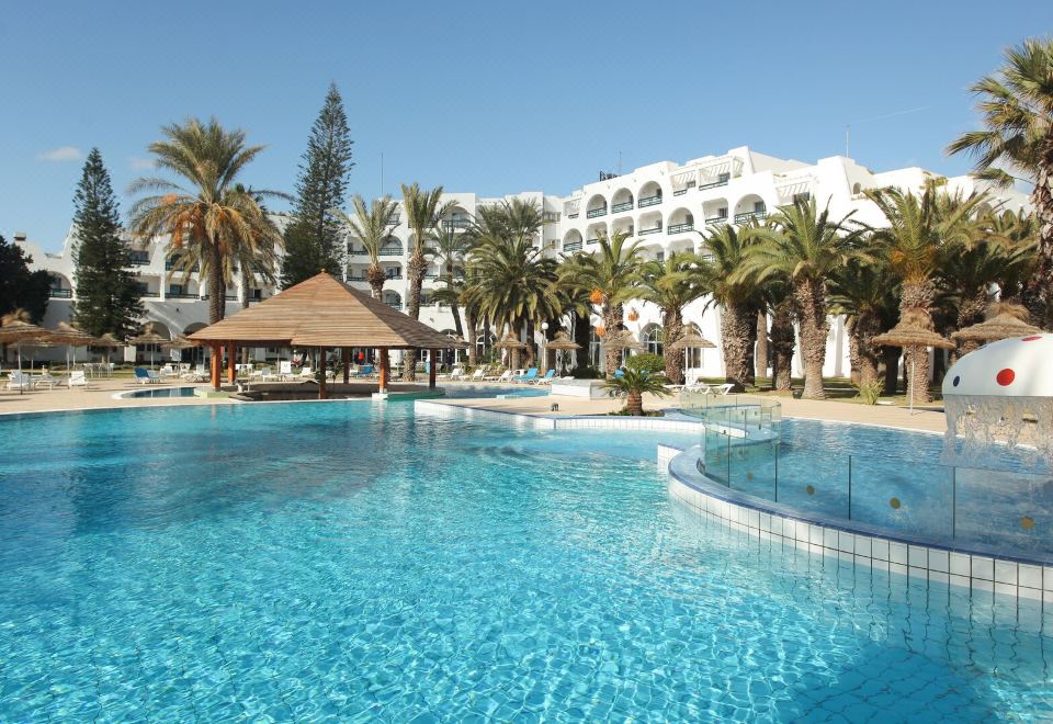 a large outdoor swimming pool surrounded by palm trees and a white building in the background at Hotel Marhaba Beach