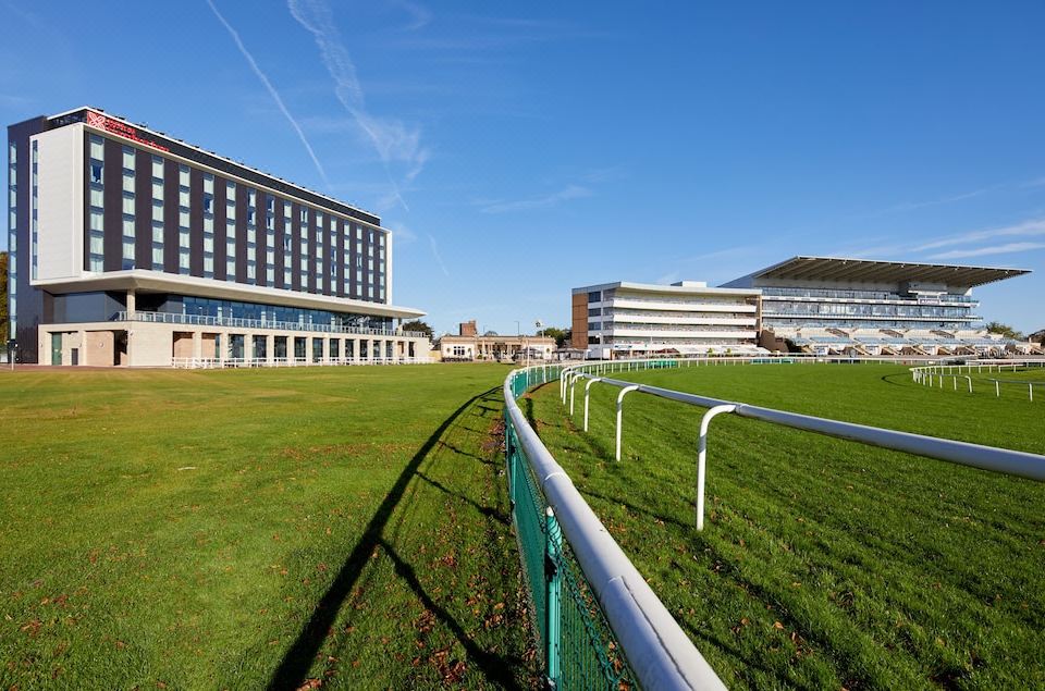 a large , modern building with a green field in front of it and a horse racing track visible in the distance at Hilton Garden Inn Doncaster Racecourse