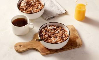 a wooden cutting board with two bowls of granola , one with milk and the other with cereal , accompanied by a glass of orange juice at Residence Inn Riverside Moreno Valley