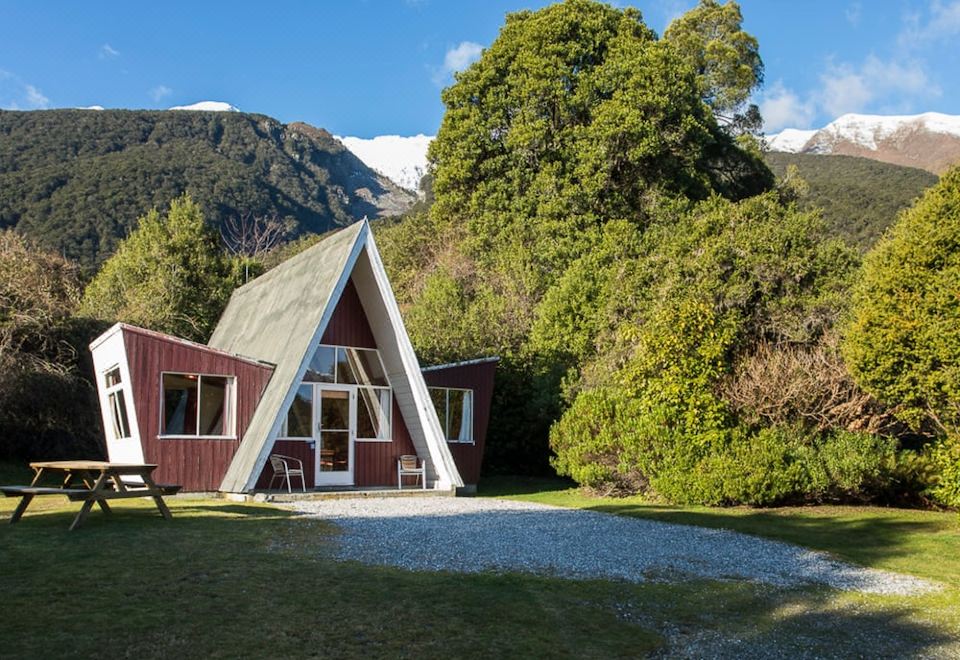 a red triangular house with a triangular roof is nestled in the mountains , surrounded by lush greenery at Wonderland Makarora Lodge