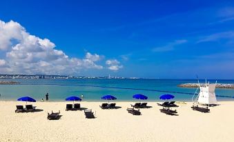 a beach scene with blue umbrellas and sun loungers , clear blue water , and a city skyline in the background at Sunset Beach Hotel