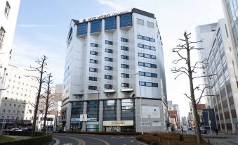 "a large white building with blue windows and a sign that says "" city hotel "" is surrounded by trees and has a road in front" at HOTEL MYSTAYS Utsunomiya
