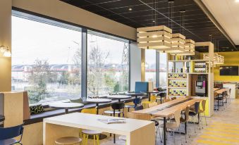 a modern , well - lit cafeteria with multiple dining tables and chairs , as well as a window offering a view of the outdoors at Ibis Styles Geneve Palexpo Aeroport