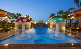 a large swimming pool with two colorful mushroom - shaped fountains in the center , surrounded by palm trees and lit up at night at Infinity See Sun Resort