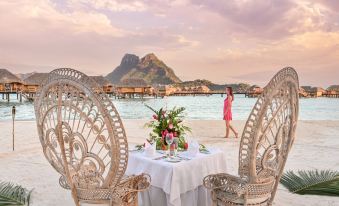 a woman in a pink dress stands near a table with two chairs and a vase of flowers on it , overlooking the ocean at Le Bora Bora by Pearl Resorts