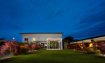 a white building with a white roof and columns is lit up at night , surrounded by green grass and bushes at Garden Hotel