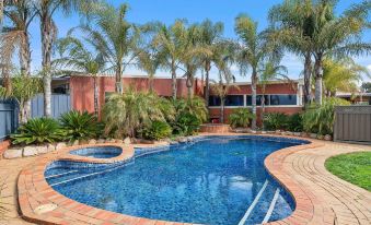 a large , kidney - shaped swimming pool surrounded by palm trees and a brick building in the background at Peppermill Inn Motel