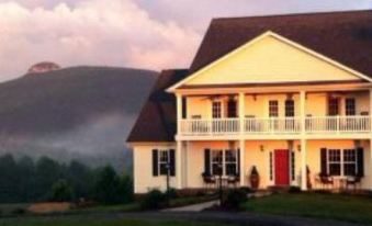 a large white house with a red door and balcony is situated on a grassy hillside , with mountains in the background at A Mighty Oak B&B
