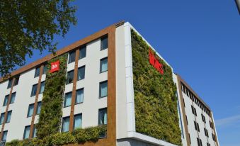 a tall building with a green wall and red sign is surrounded by trees and other buildings at ibis Lyon Sud Oullins