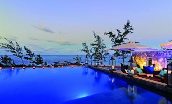 a large , blue swimming pool surrounded by palm trees and a wooden deck , with the ocean visible in the background at Constance Tekoma