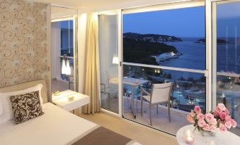a bedroom with a bed and a balcony overlooking a pool at night , creating a serene atmosphere at Amfora Hvar Grand Beach Resort