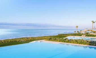 a large swimming pool is surrounded by grass and trees , with the ocean visible in the background at Ikos Andalusia