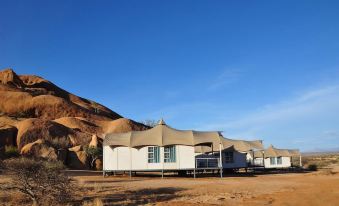 a row of beige and white tents is set up in a desert area , surrounded by mountains at Spitzkoppen Lodge