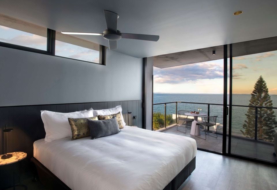 a large bed with white linens is in a room with a sliding glass door that leads to an ocean view at The Sebel Brisbane Margate Beach