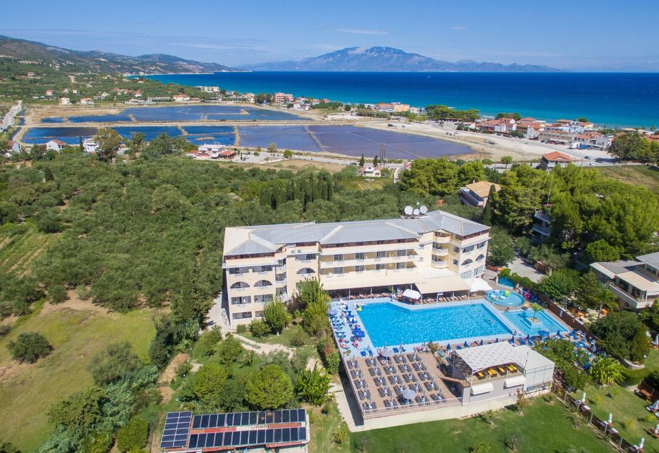 aerial view of a resort with a large pool surrounded by buildings , a golf course , and a body of water in the background at Koukounaria Hotel & Suites