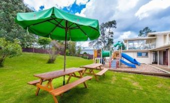 a large green umbrella is set up over a picnic table with benches in a grassy area near a playground at Poenamo Hotel