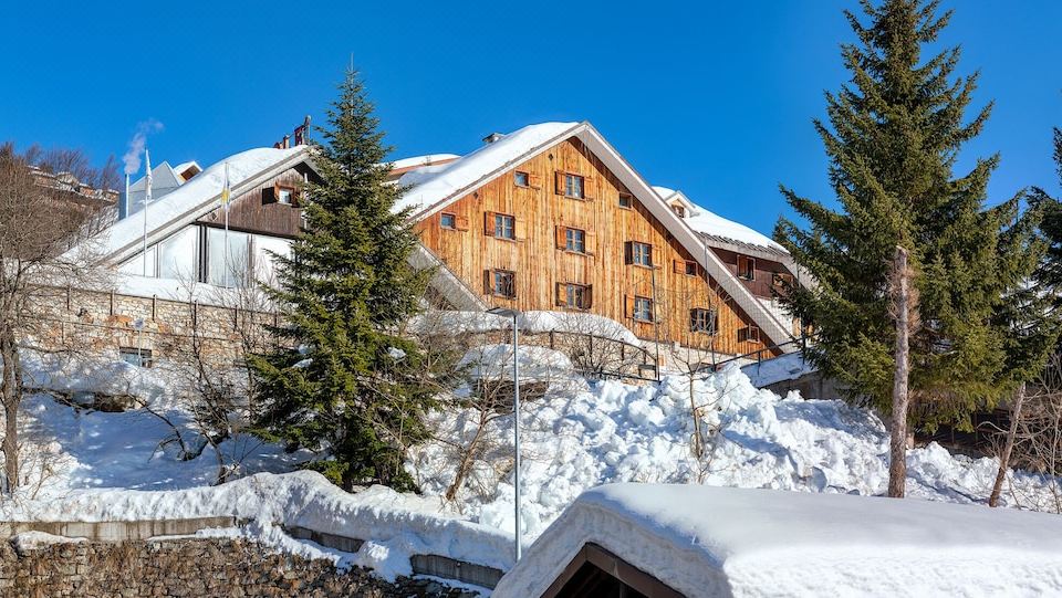 a wooden building with a sloping roof is perched on a snow - covered hill , surrounded by trees and clear skies at Hotel Mondole