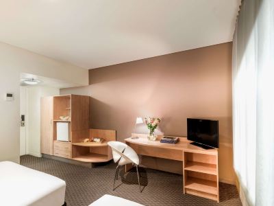 a modern hotel room with a desk , chair , and tv , set against a beige wall at Ibis Melbourne Hotel and Apartments