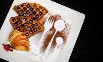 a white plate with a waffle topped with apples and a chocolate sauce , accompanied by a fork at Hotel Avenue