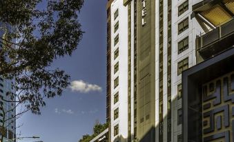"a tall building with a red sign that reads "" ibis "" is surrounded by trees and other buildings" at Ibis Melbourne Hotel and Apartments
