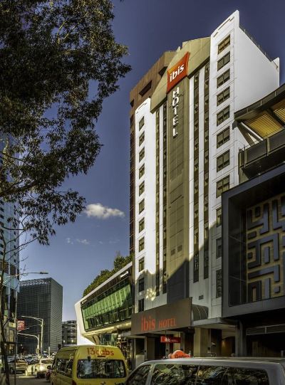 a modern city street with a tall building and a hotel on the corner , surrounded by trees at Ibis Melbourne Hotel and Apartments