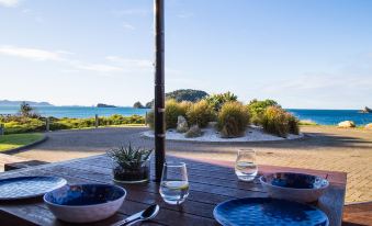 a table with plates , wine glasses , and a bowl of food is set up on a deck overlooking the ocean at Hahei Beach Resort