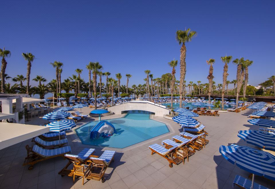 a large outdoor pool surrounded by lounge chairs and umbrellas , with palm trees in the background at GrandResort by Leonardo Hotels