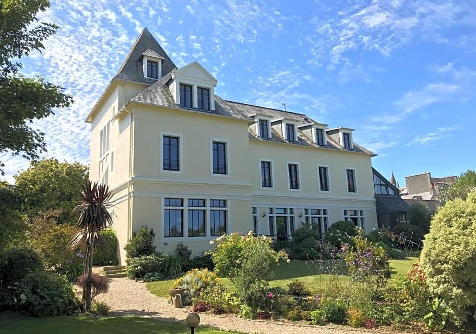 a large , yellow building with a triangular roof and blue windows is surrounded by a lush garden at Hotel de France